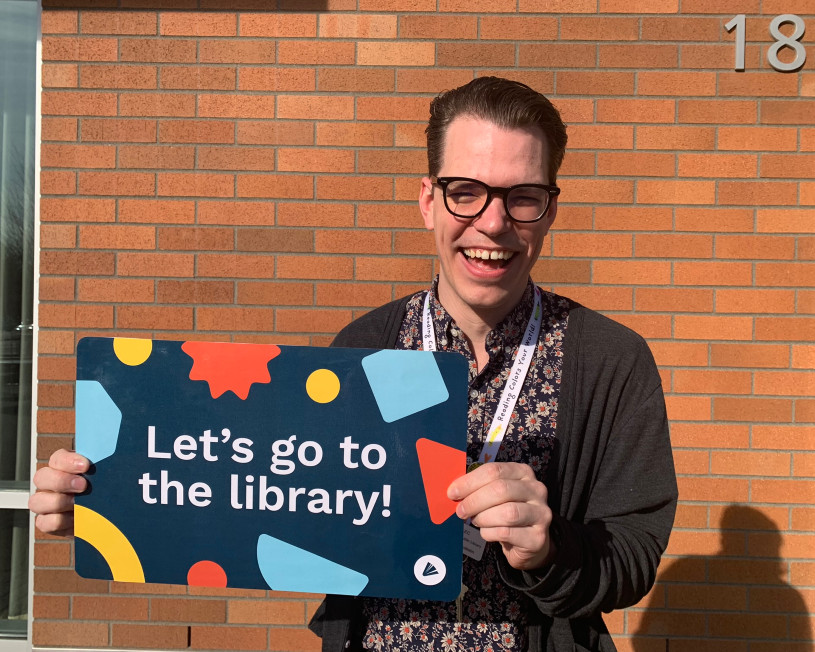 Bibliotecario sosteniendo una tarjeta de biblioteca gigante que dice: ¡Vamos a la biblioteca!