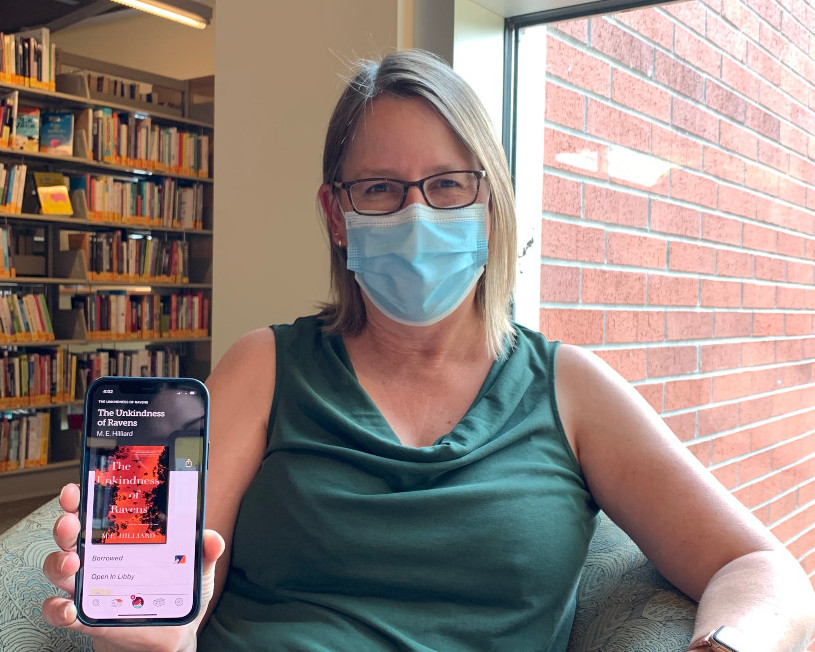 Woman wearing a disposable mask and glasses sits by the window in a library reading on her phone.