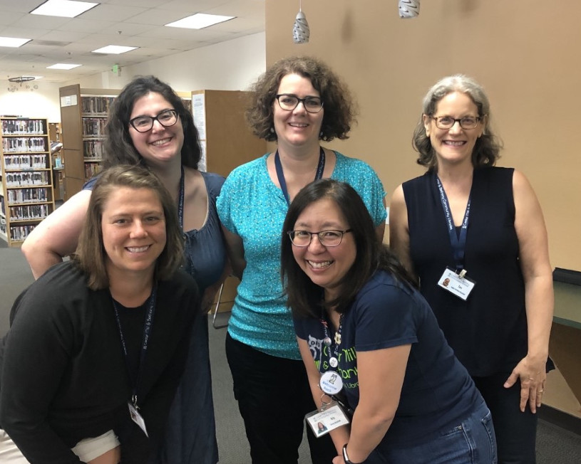 Five women gather round in the library and smile at the camera