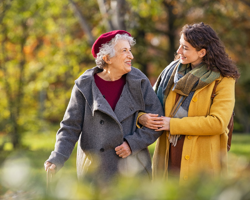 Una mujer mayor con un bastón y una mujer más joven caminan del brazo afuera en un día de otoño.