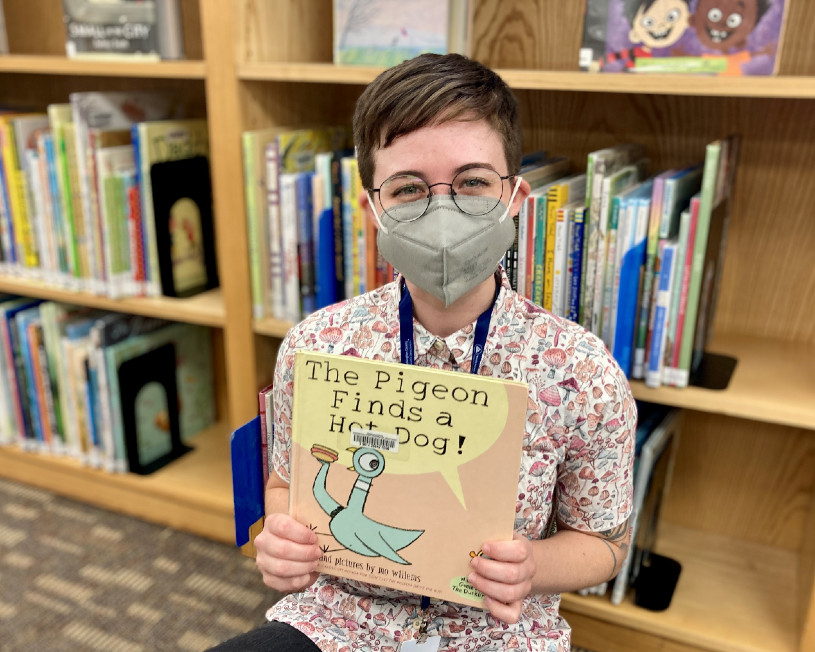 librarian holding the book "The Pigeon Finds a Hot Dog" sitting in front of a bookshelf