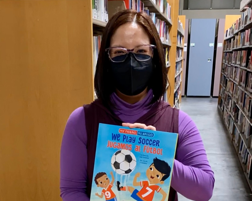 Woman wearing glasses and a mask, holds up a children's book about soccer.