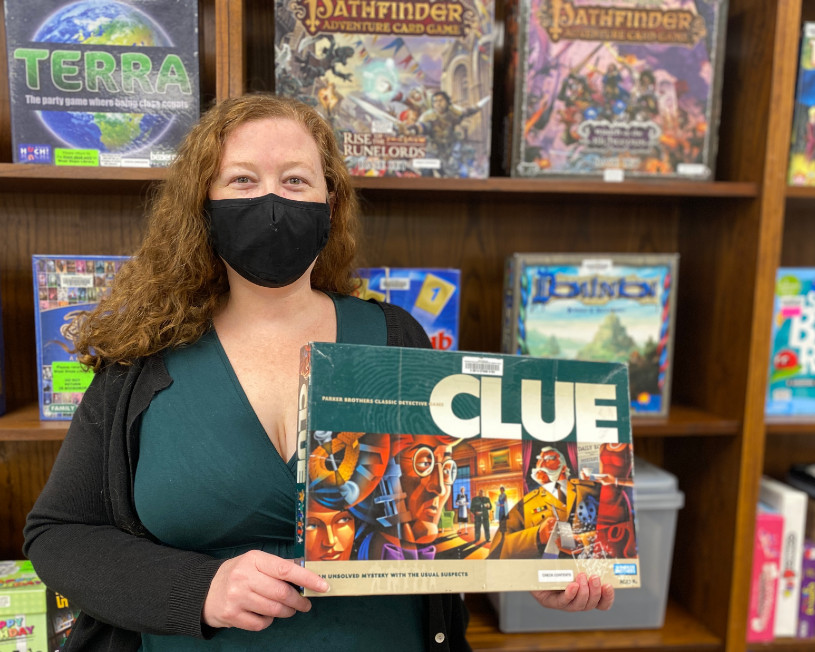 Woman wearing a green dress and a mask holds up the board game Clue in front of a game display.
