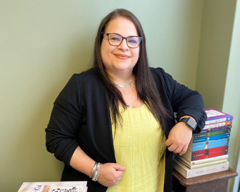 Woman wearing glasses stands next to stacks of young adult novels in English and Spanish