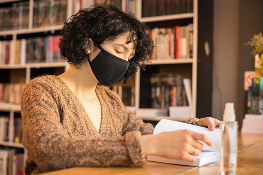Mujer enmascarada mirando un libro en la biblioteca, desinfectante de manos en la mesa