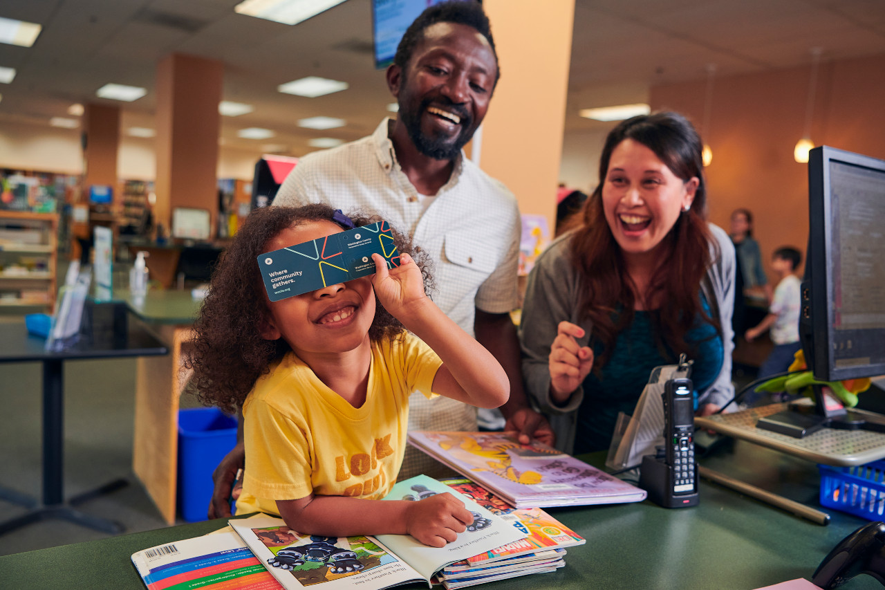 A child holding a WCCLS library card and two laughing adults looking on