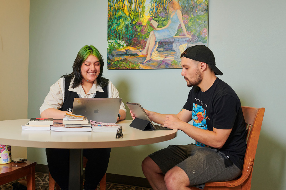 Woman with laptop and man with tablet, seated at a table with books