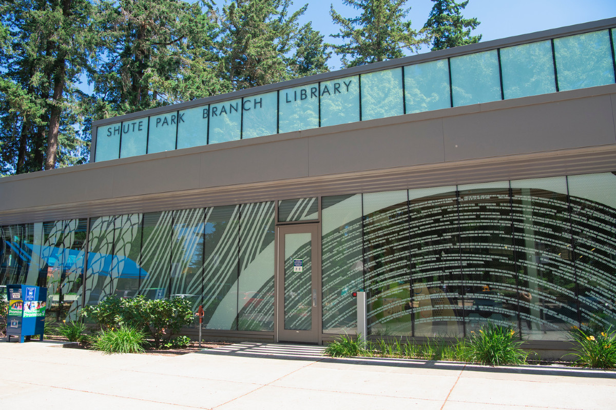 Pared exterior de vidrio decorada con líneas curvas de texto, etiquetada como "Biblioteca sucursal de Shute Park" en la parte superior izquierda