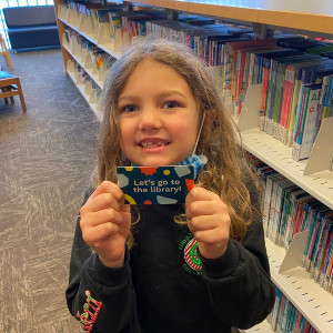 Niña sosteniendo su tarjeta de la biblioteca y sonriendo en la sección infantil de la biblioteca