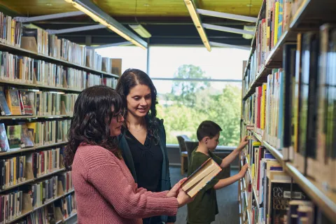 Two women and young boy looking at books on a shelf in the library.