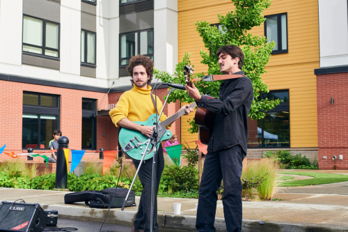 Two men play guitar outside a brick-faced building with greenery