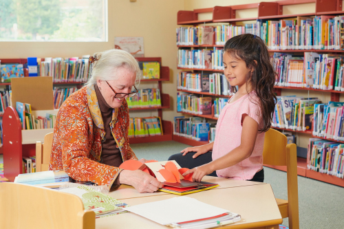 Mujer de cabello blanco y niña miran un libro emergente en una mesa en una biblioteca