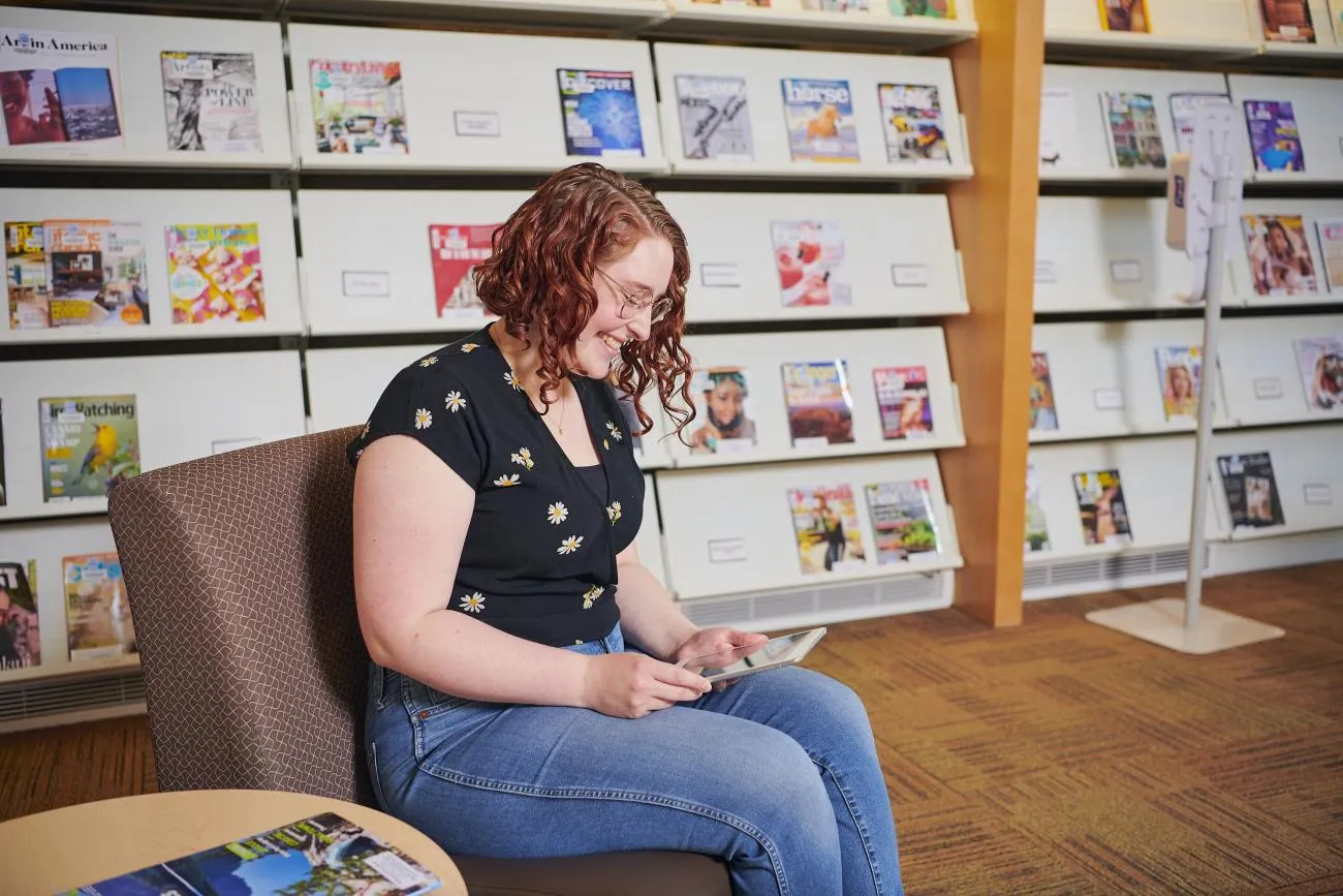 Una persona con cabello rizado rojo y gafas lee en una tableta frente a un expositor de revistas.