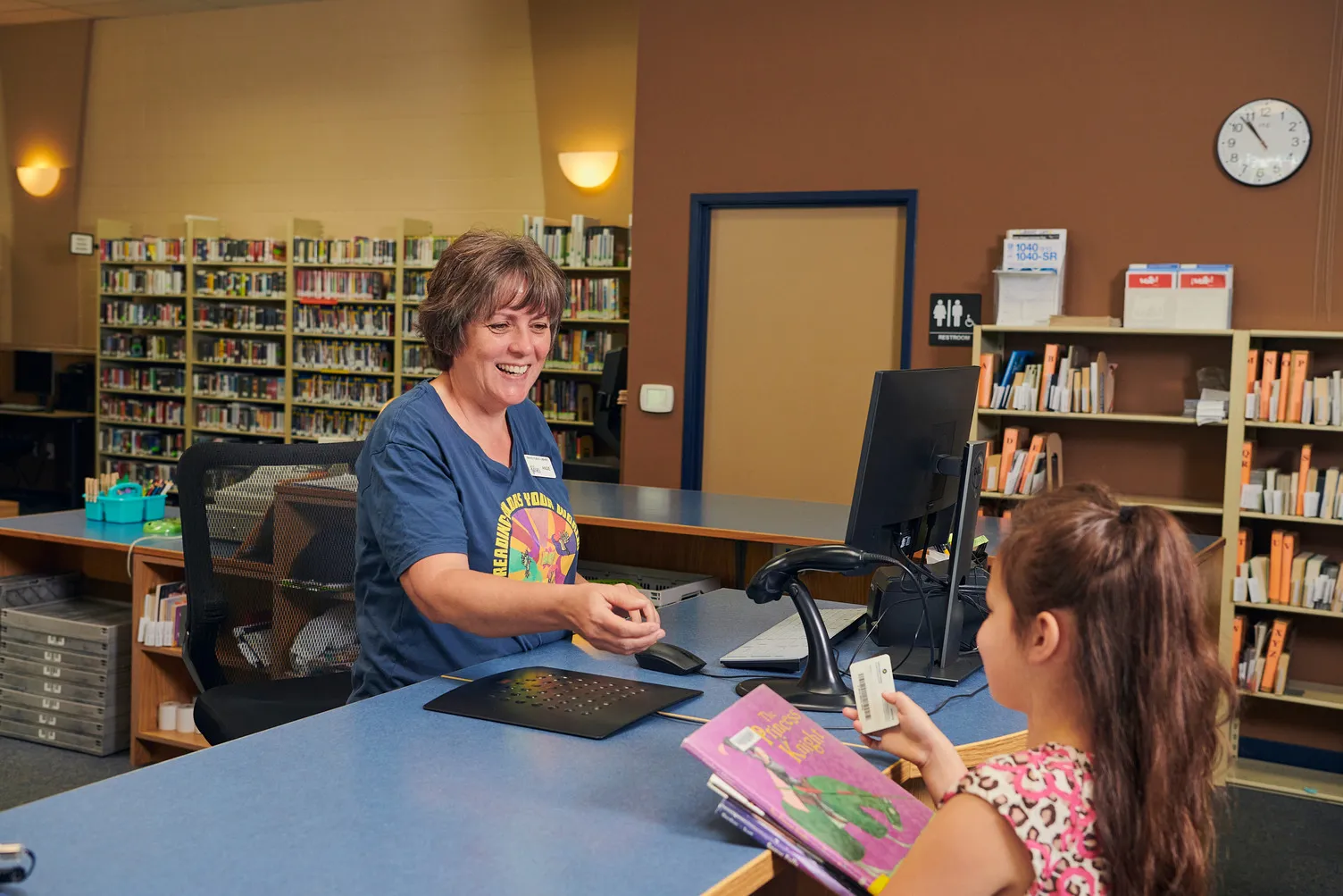 Library staff smiling and reaching over desk to grab a library card from a young girl. The young girl is holding her library card in her right hand and a stack of picture books in her left hand.