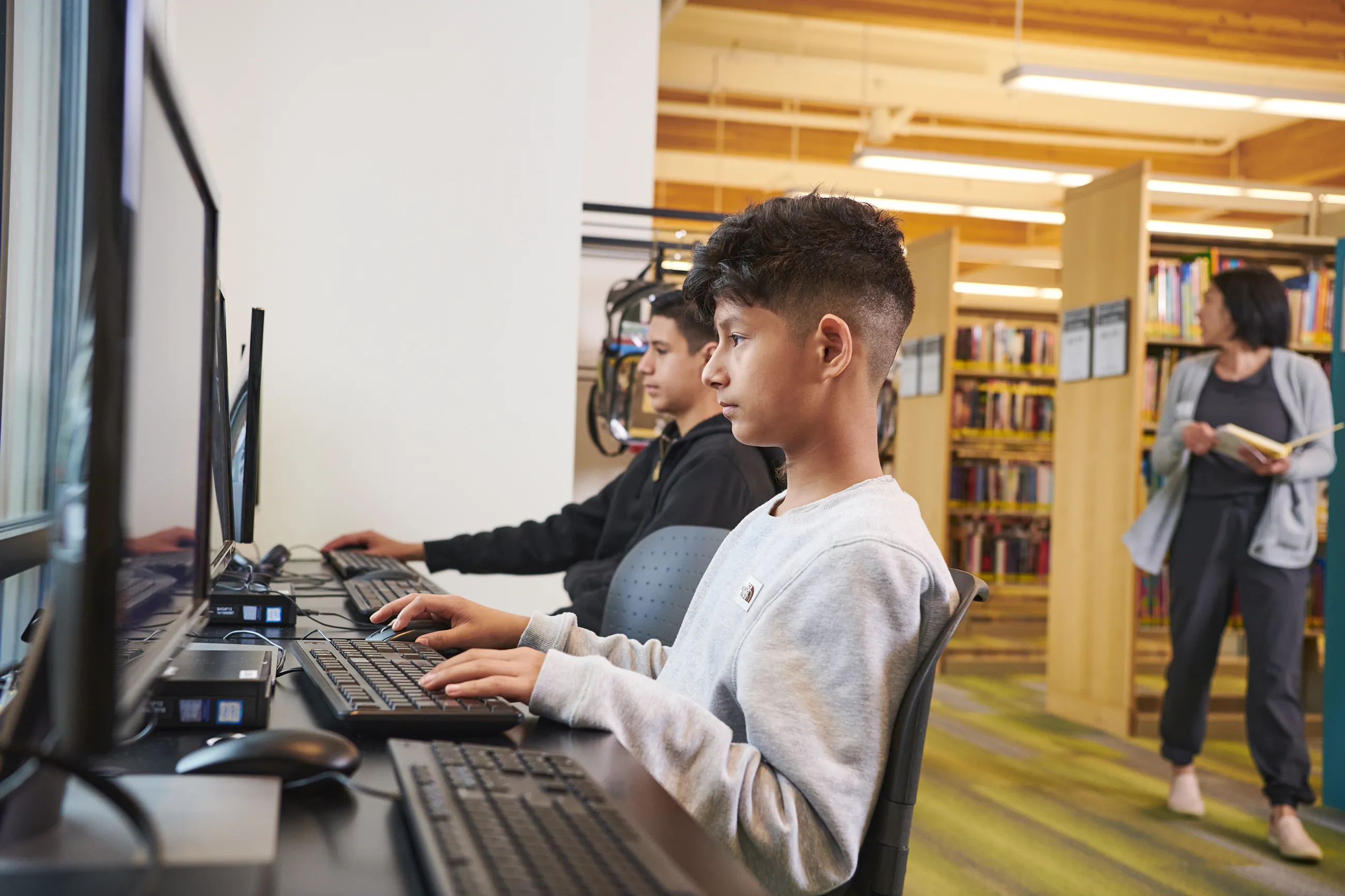 Two teen boys are sitting at desktop computers in the library.