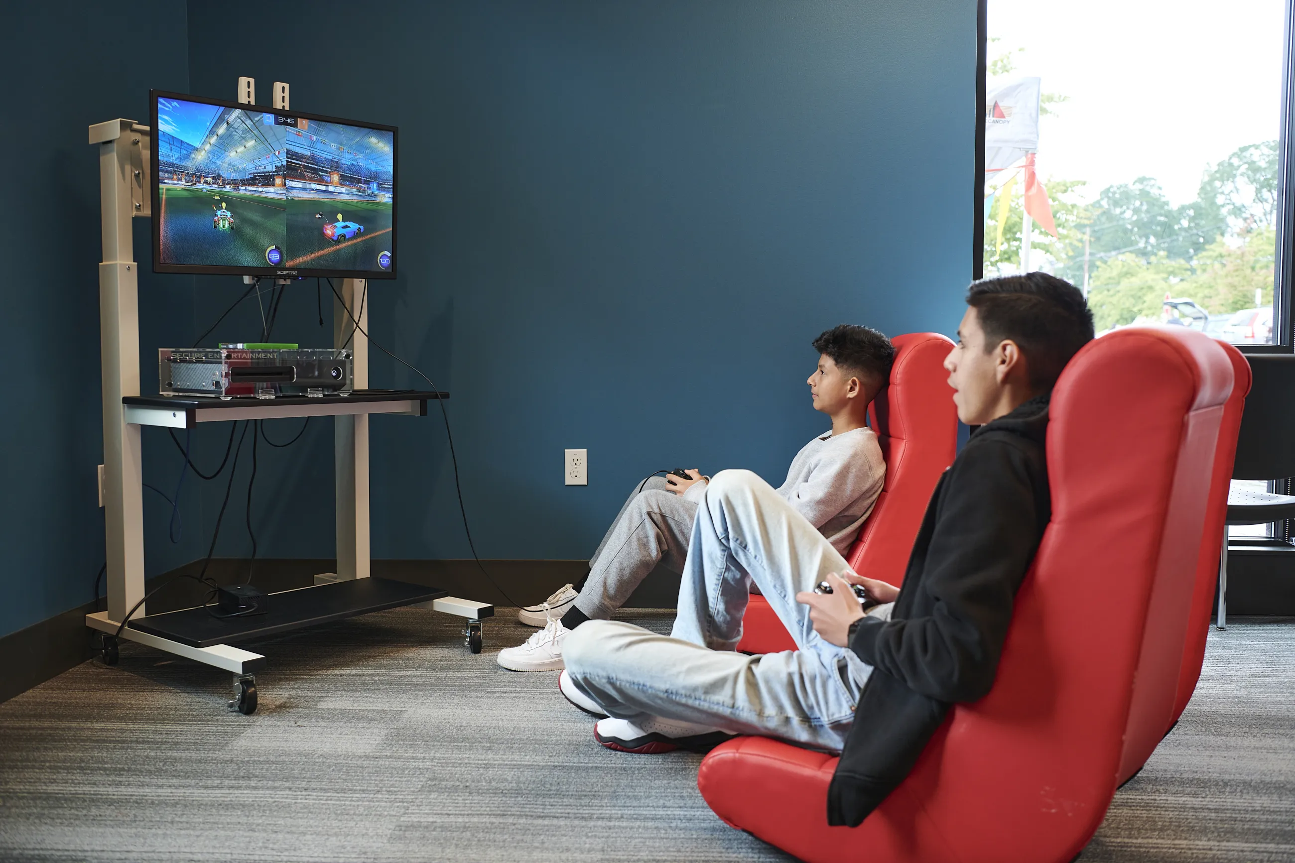 Two teen boys sitting in gaming chairs and looking at a tv while playing a video game.