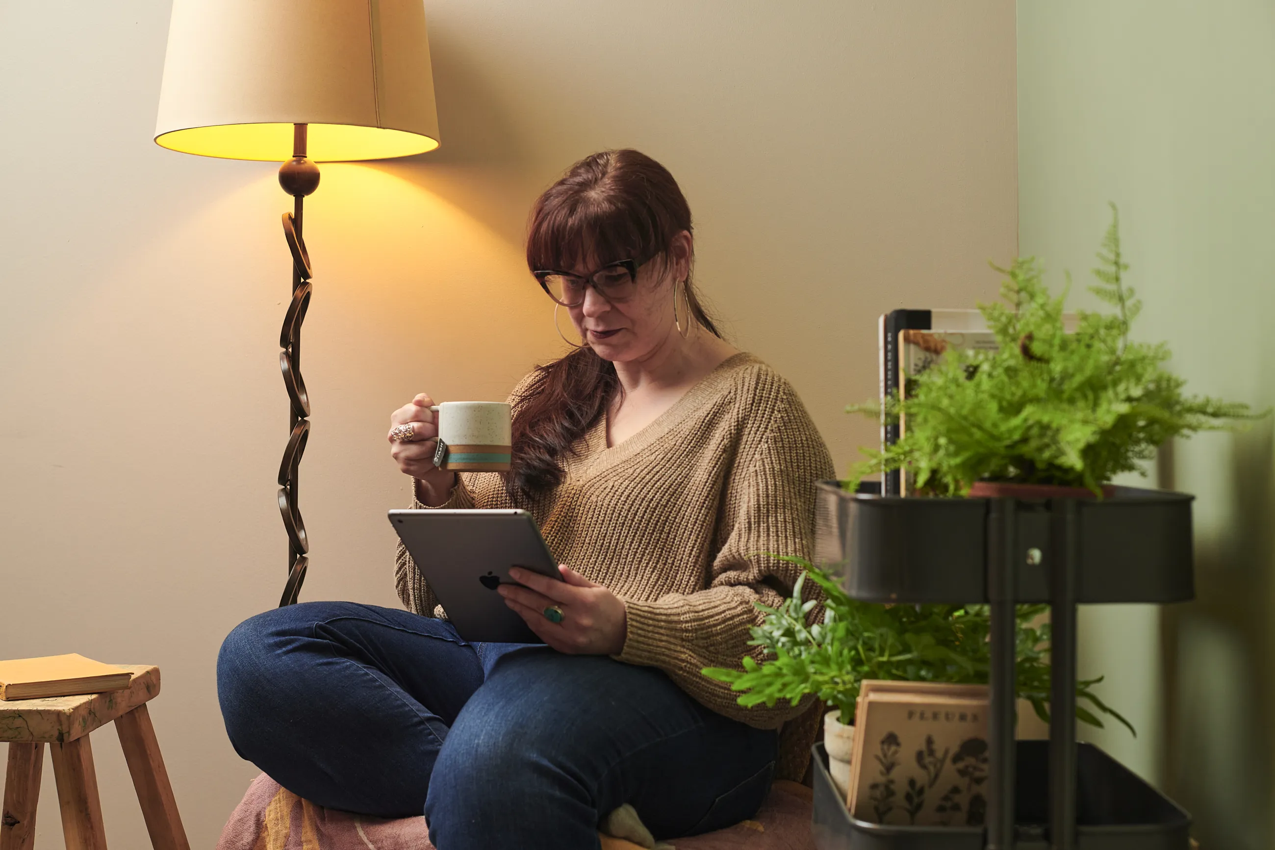 Woman with a long dark ponytail and glasses reads on a tablet while holding a mug