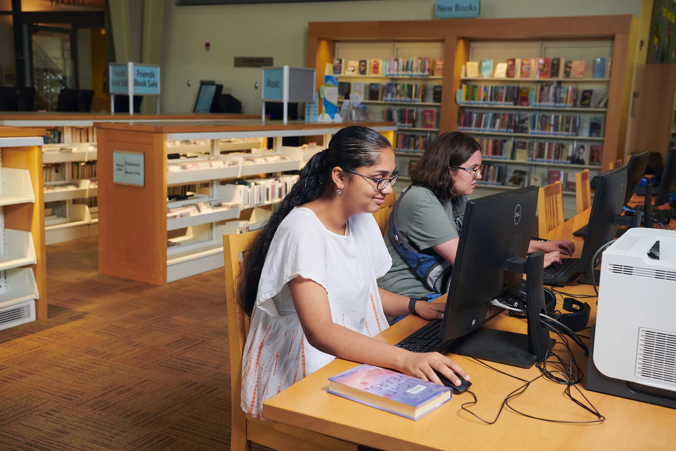 Teen girl with long black hair and wearing white top is sitting at a desktop computer in the library.