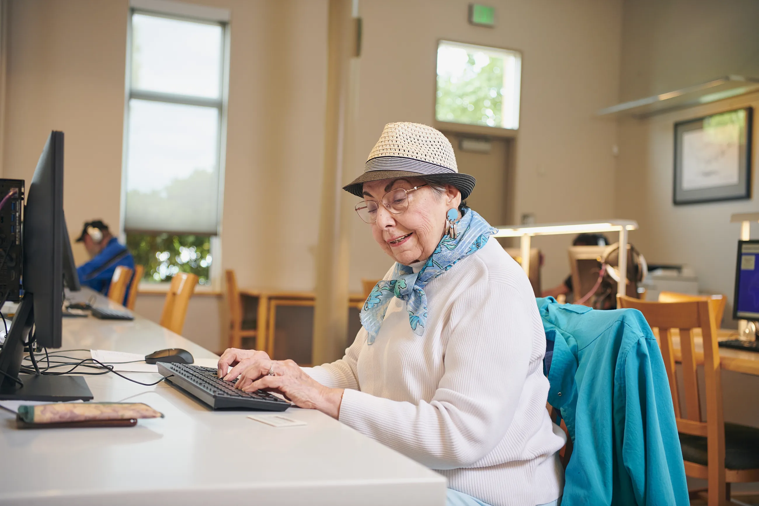Person with hat and glasses and a blue scarf types on a computer at the library