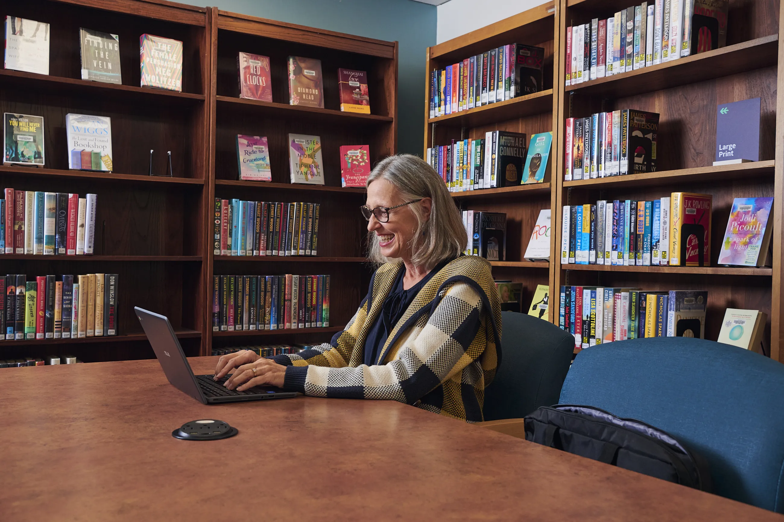 Person with glasses and plaid coat sits at a table in the library, using a laptop