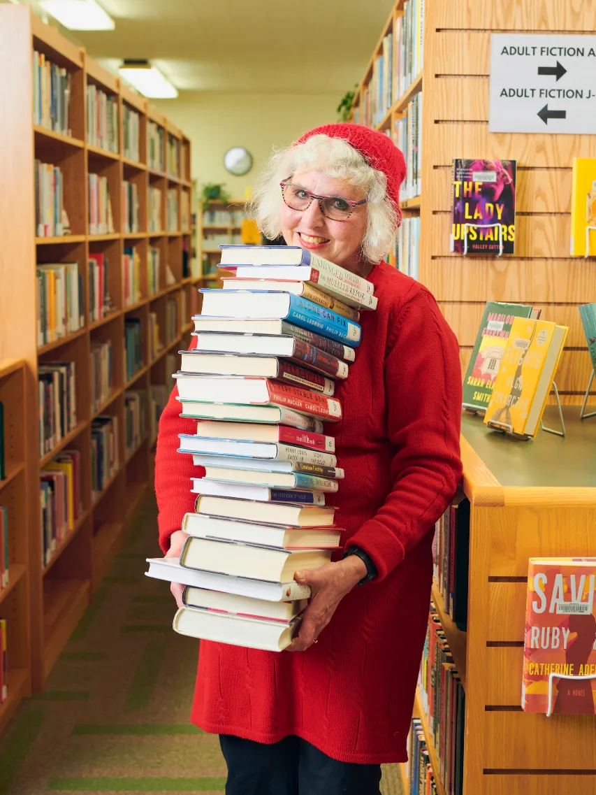 Woman wearing red dress and red hat holding a tall stack of books in front of bookshelves.
