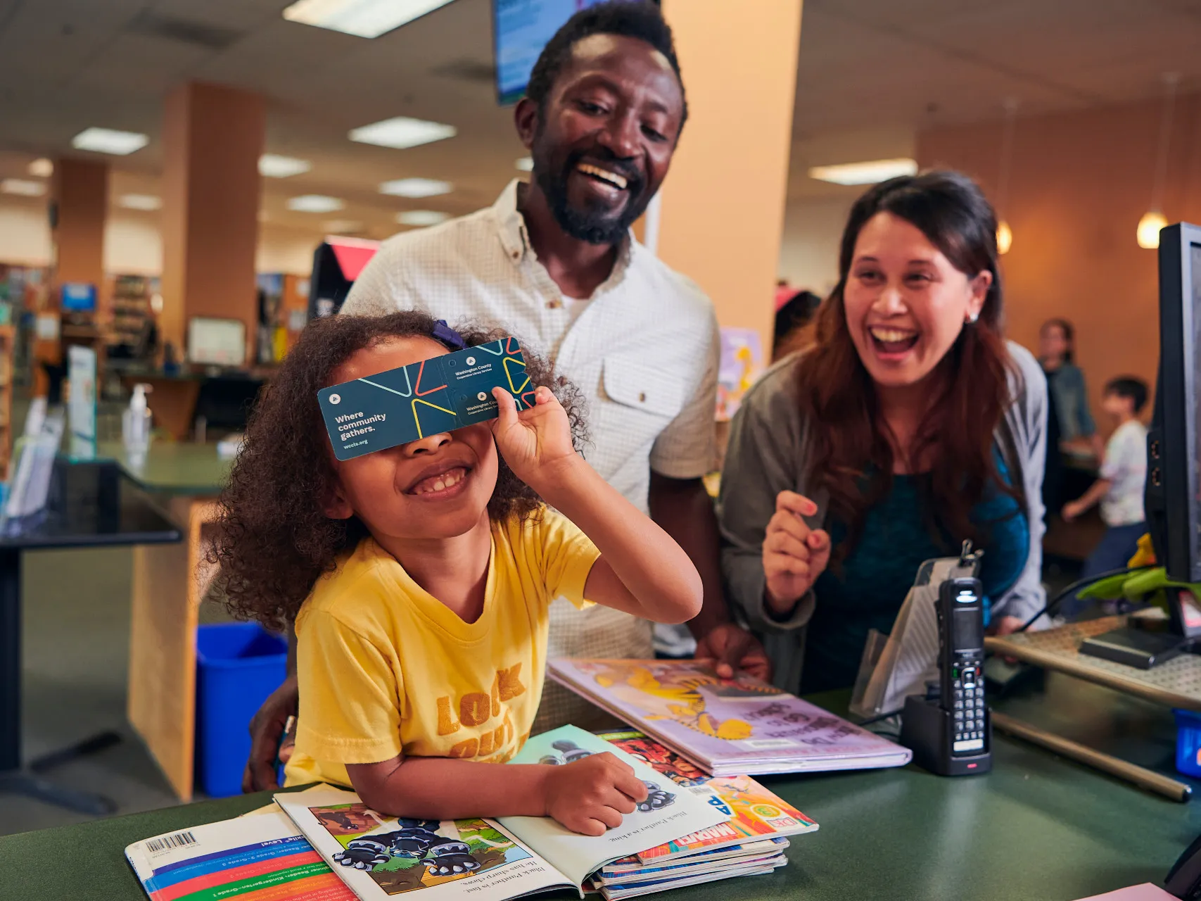 Smiling kid in yellow shirt covers eyes with a library card while parents giggle nearby