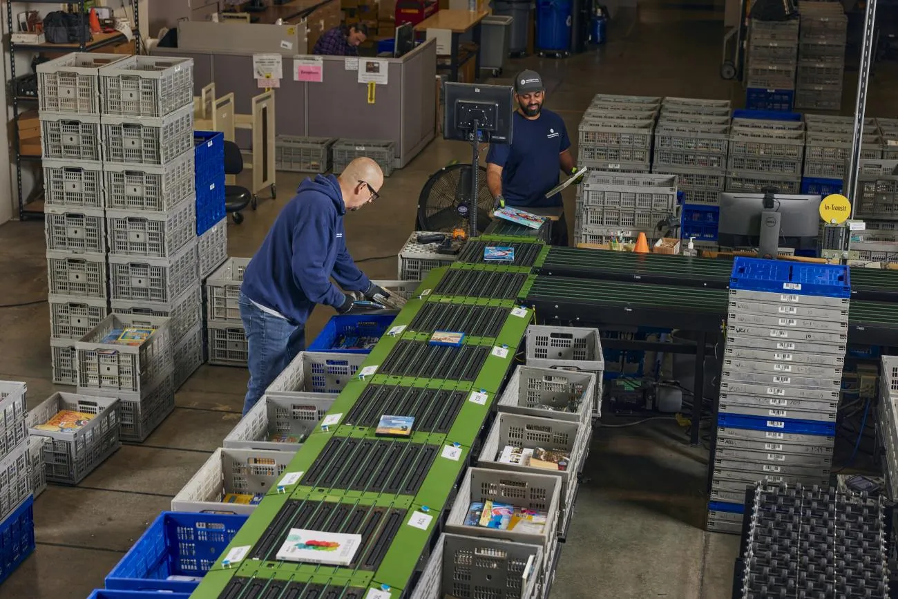 two men working at conveyer belts and holding books in a warehouse