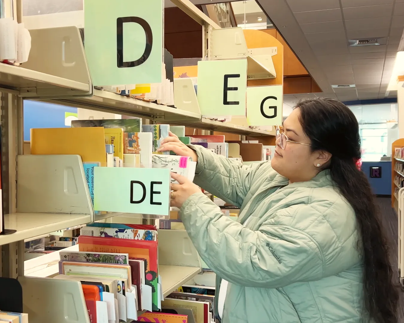 Woman with long black hair and wearing green jacket is taking a book off of the holds shelf in the library.