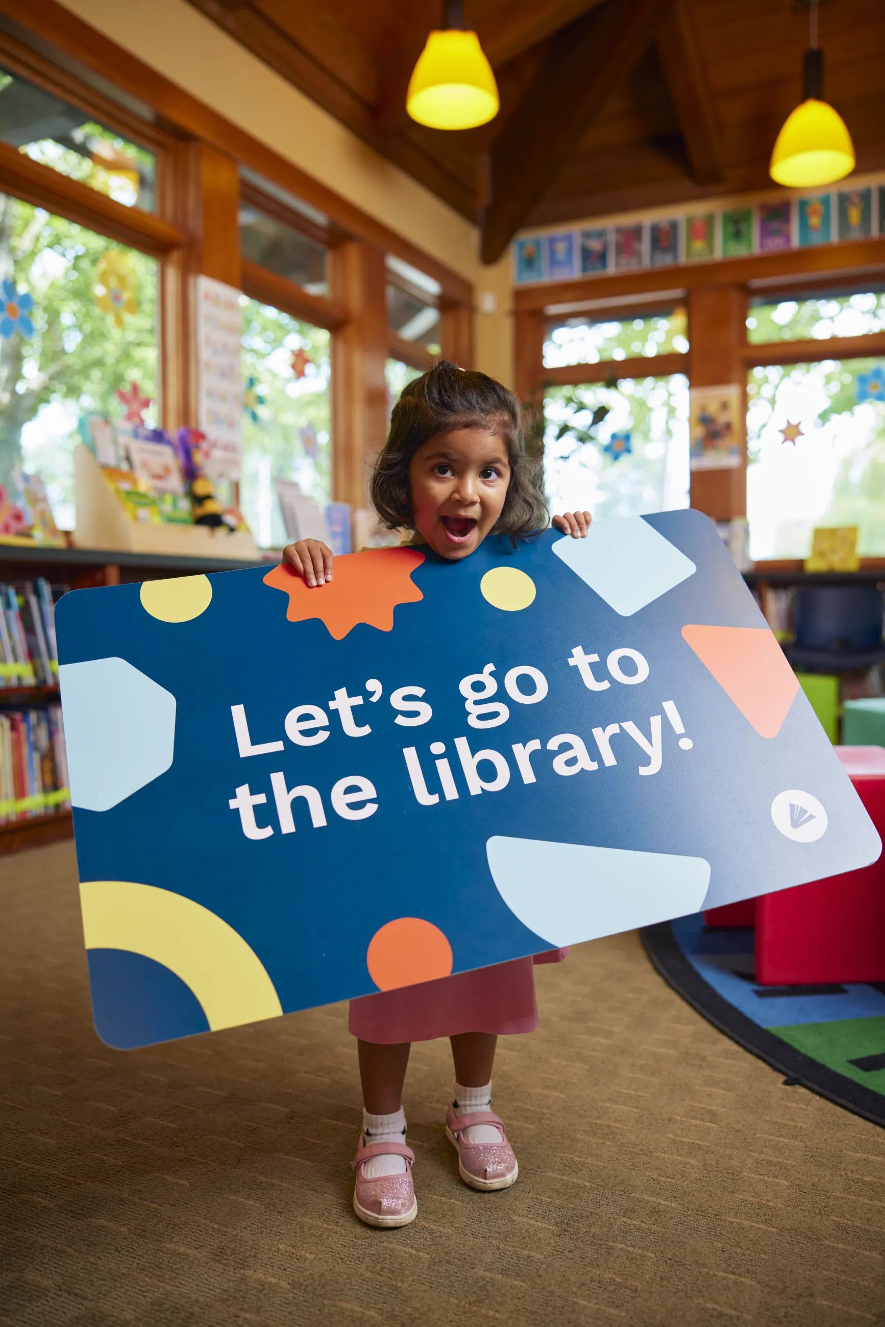Young girl with short brown hair and wearing a pink dress and pink shoes is holding a giant WCCLS library card with colorful shapes on it and the words "Let's go to the library!".