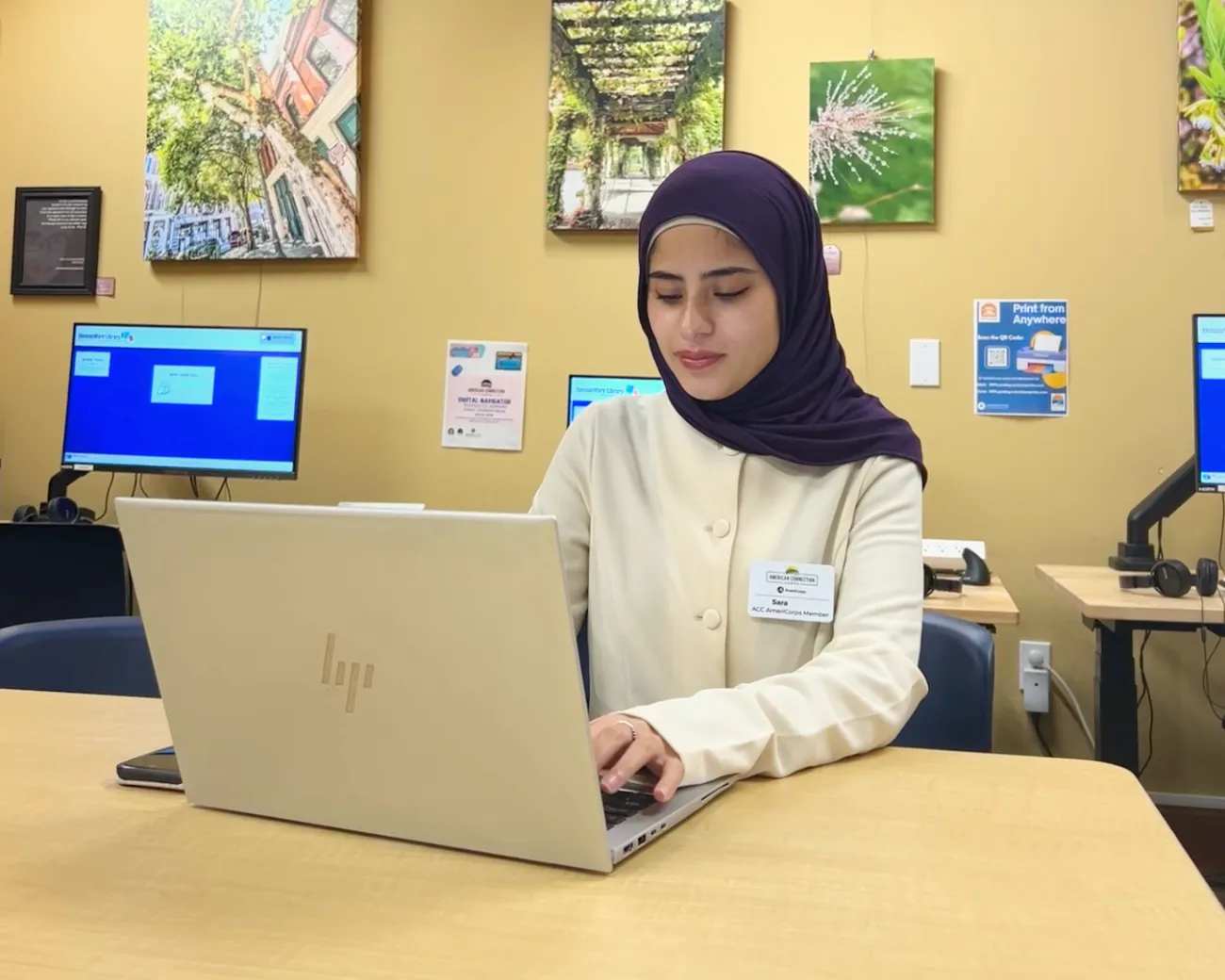 A woman works on her laptop at a table, with computer stations behind her.