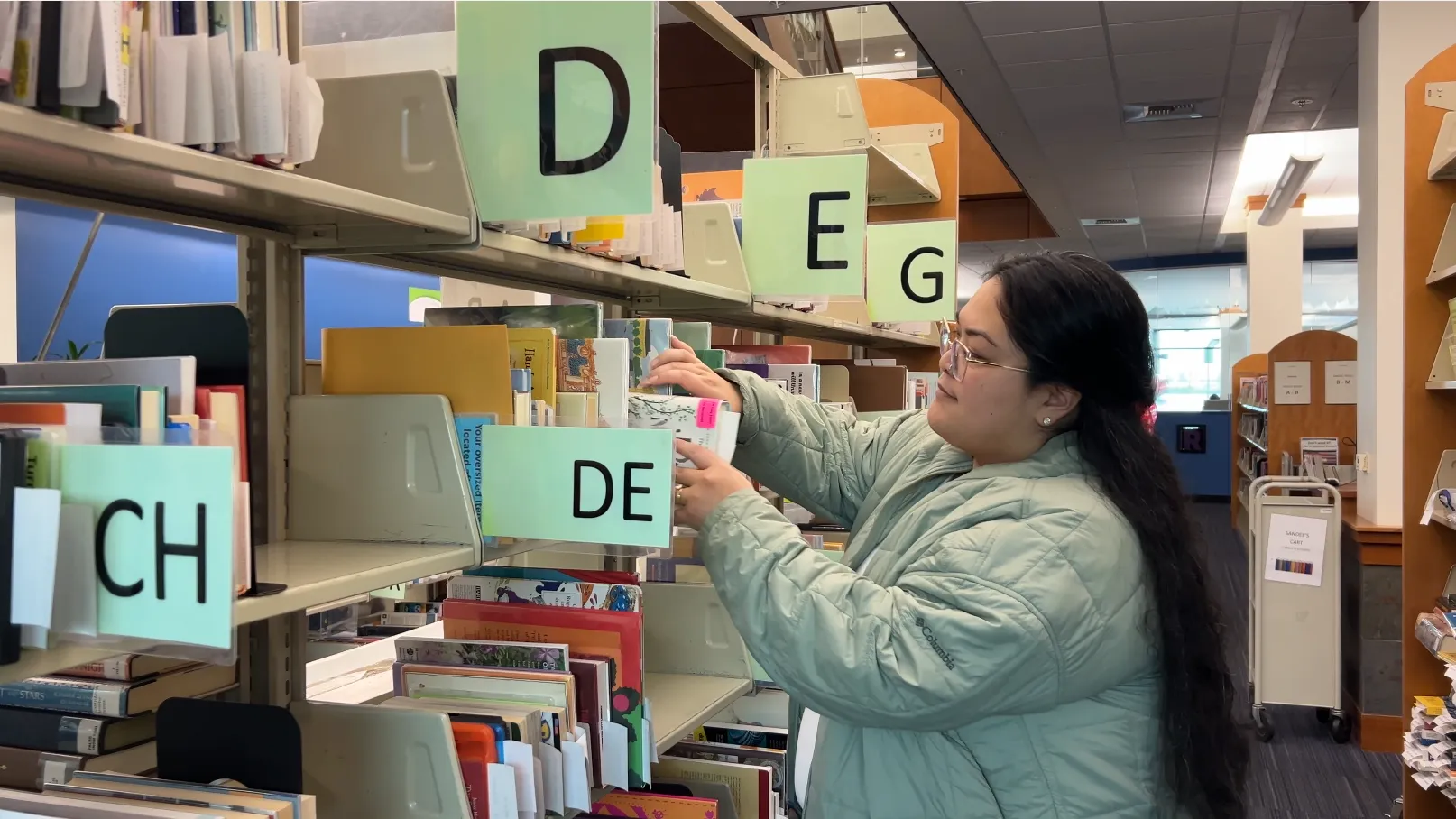 Woman long black hair and wearing green jacket is pulling a book off of the shelf at the library.