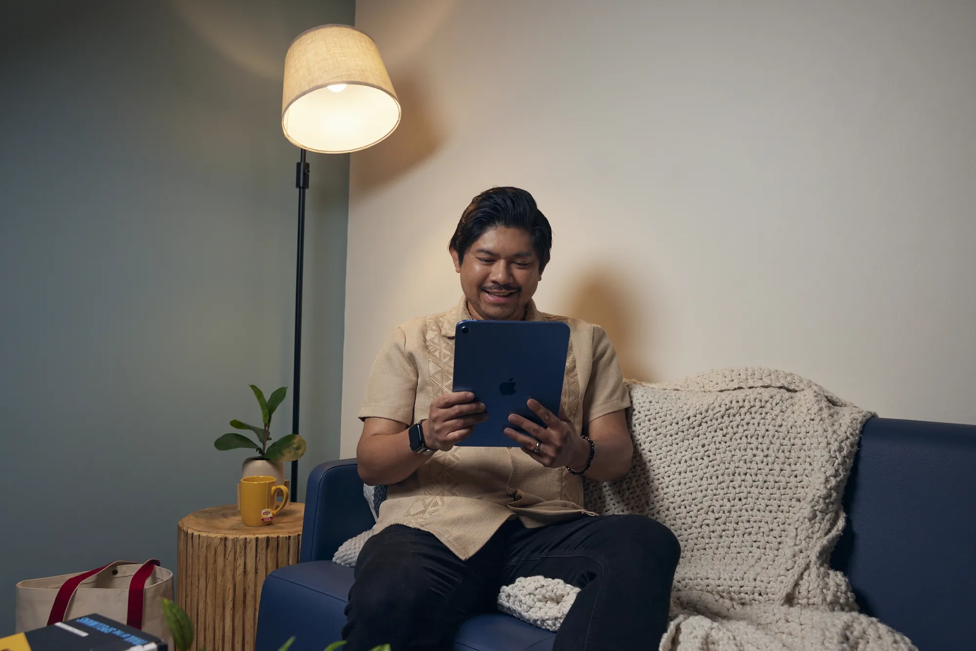 Man wearing tan short sleeve shirt and sitting on couch. He is looking at the screen of an iPad while smiling.