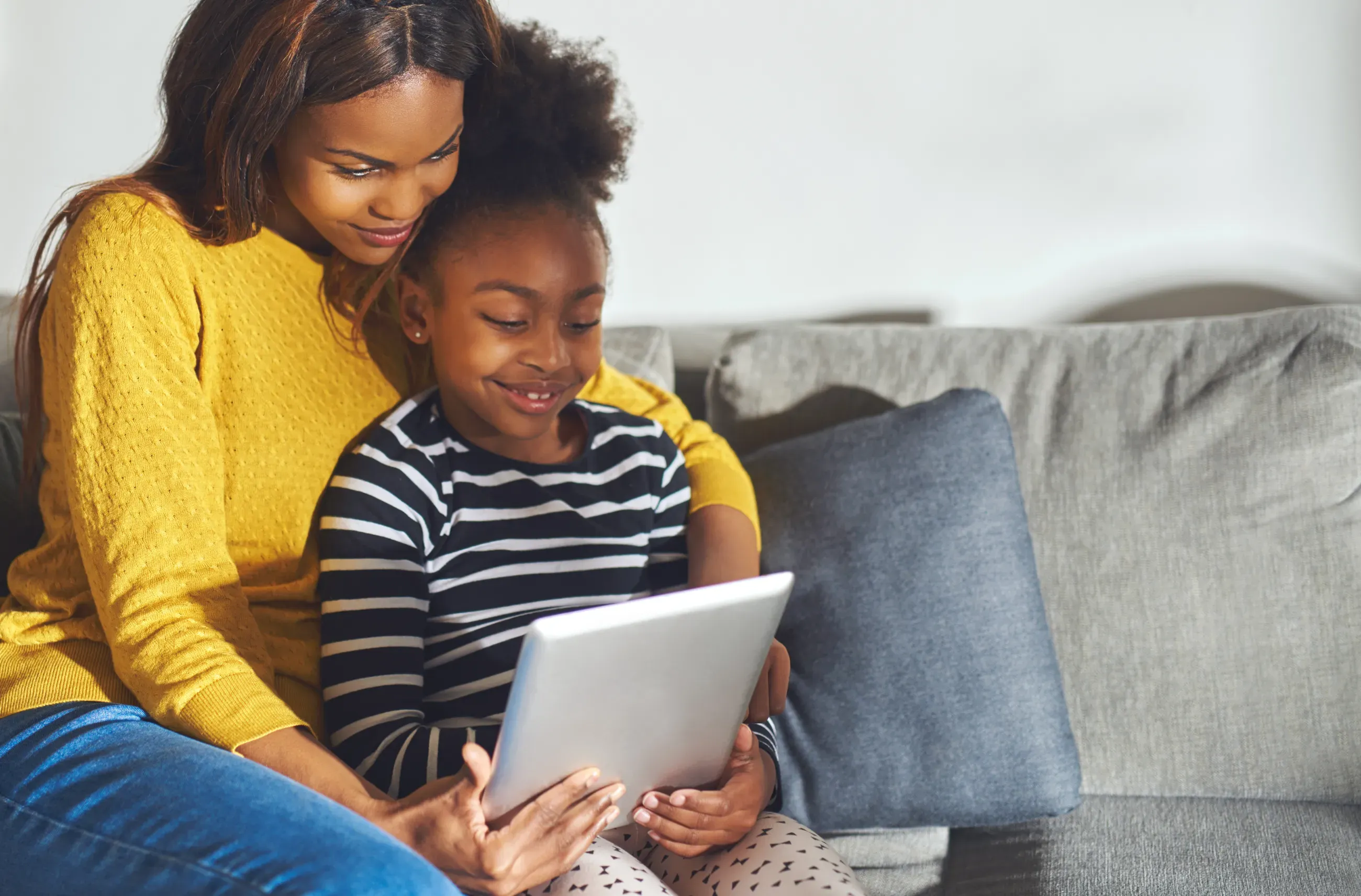 A mom and daughter sitting on a couch reading a tablet together.