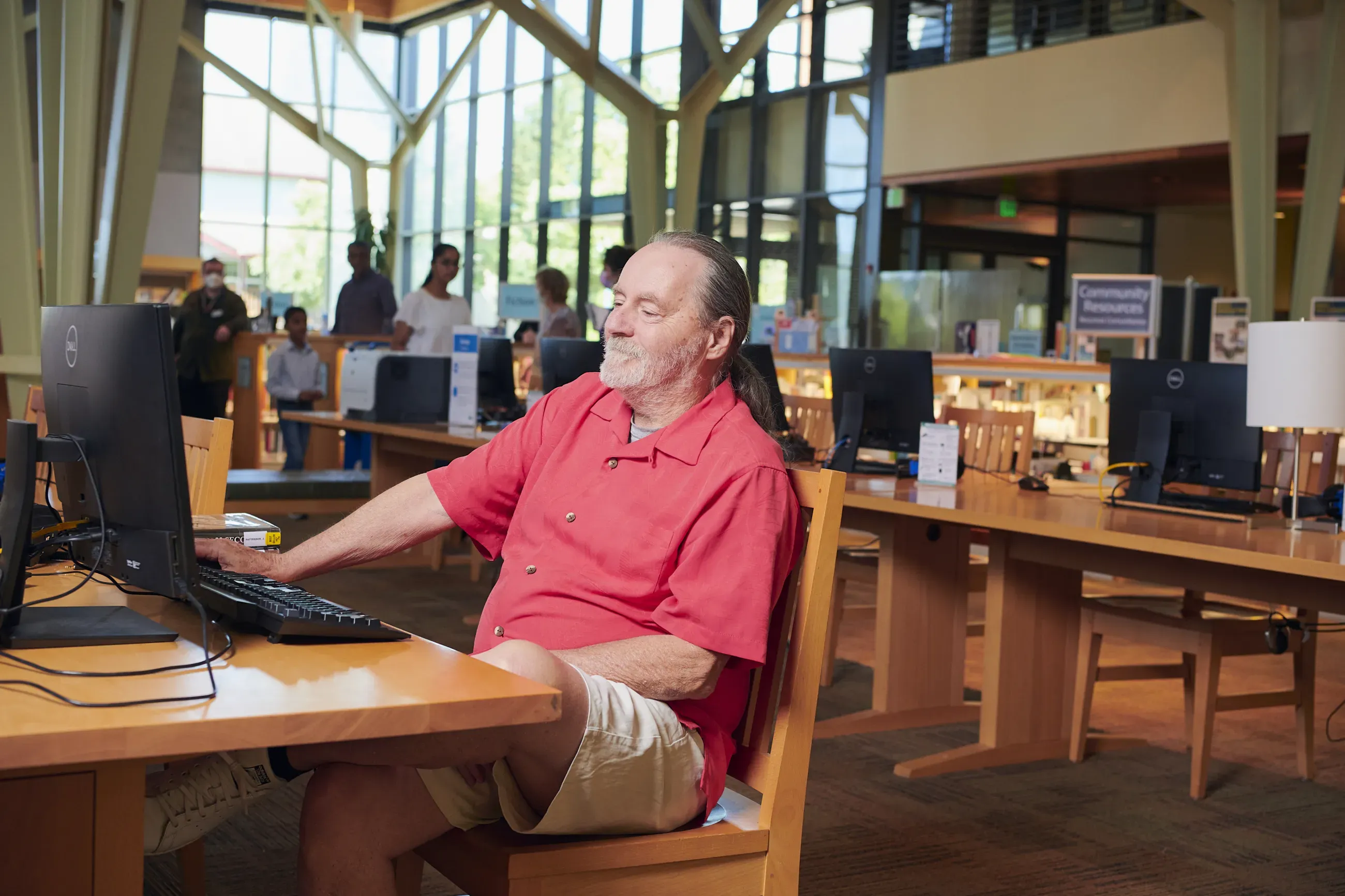 Man in a red shirt sitting at a computer in a library with many windows.