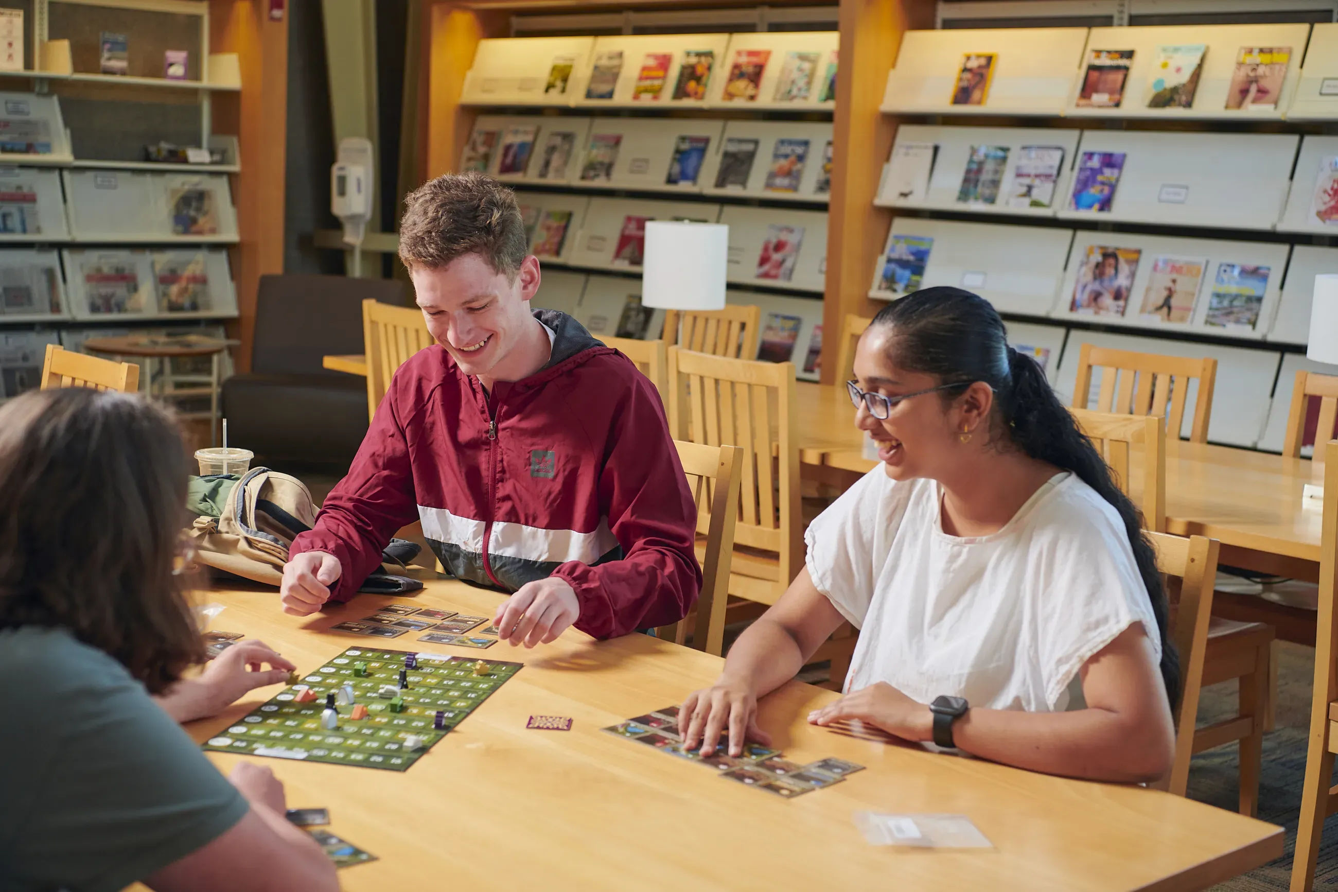 Three young adults play a board game at a table in a library.