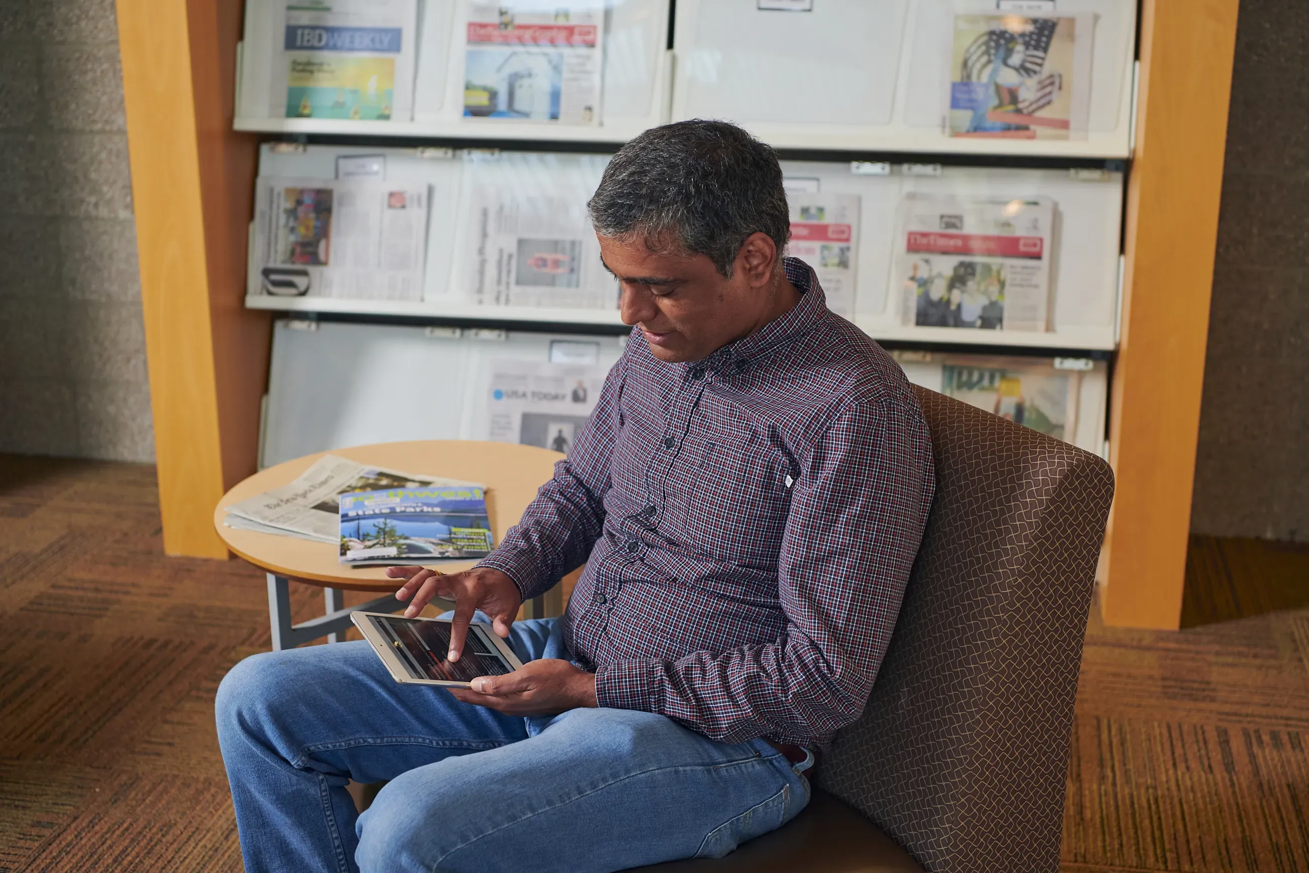 Person wearing button-down shirt and jeans scrolls on a tablet while sitting in a library.
