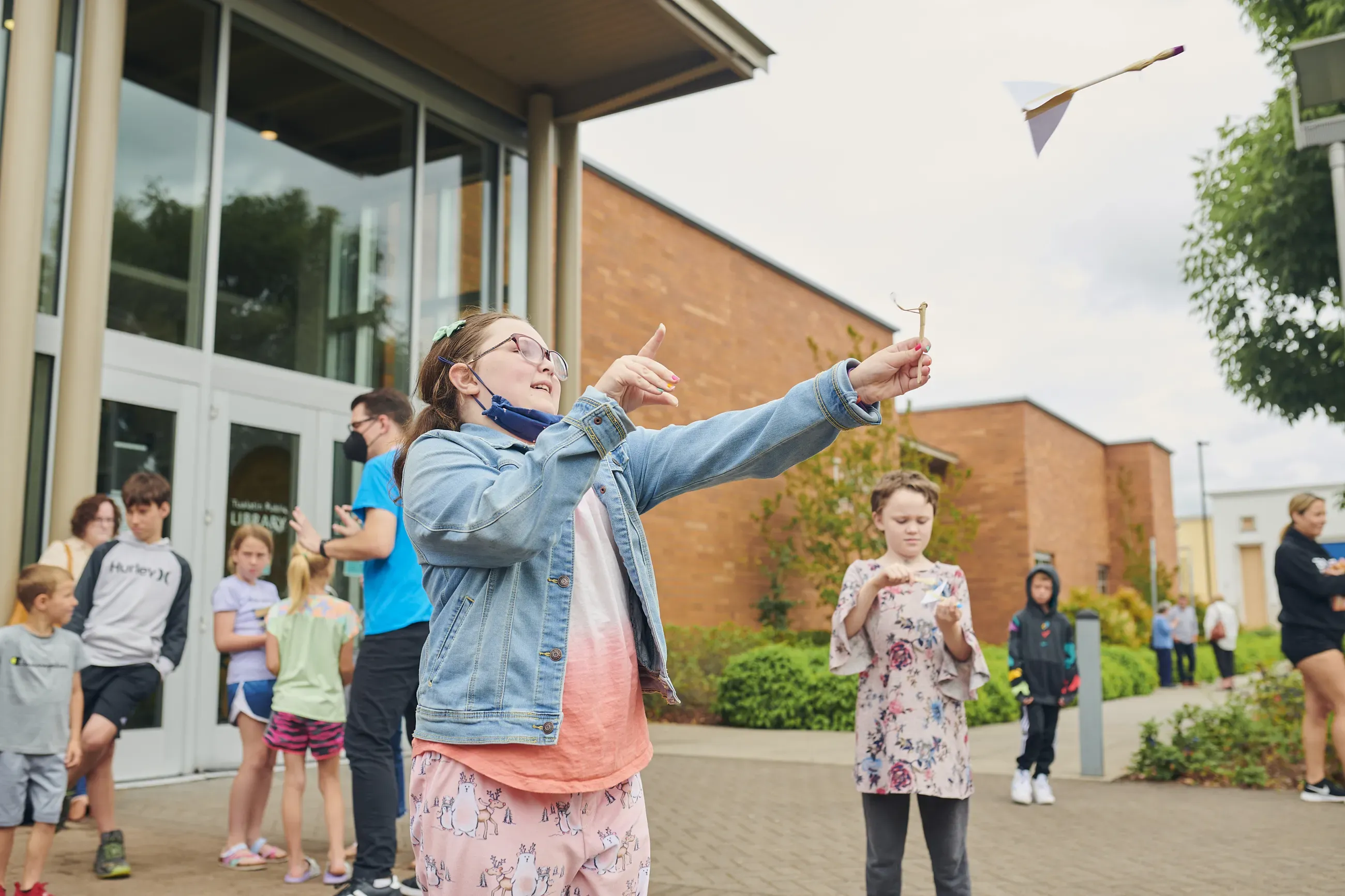 Children outside a library, and one girl launches a paper rocket.