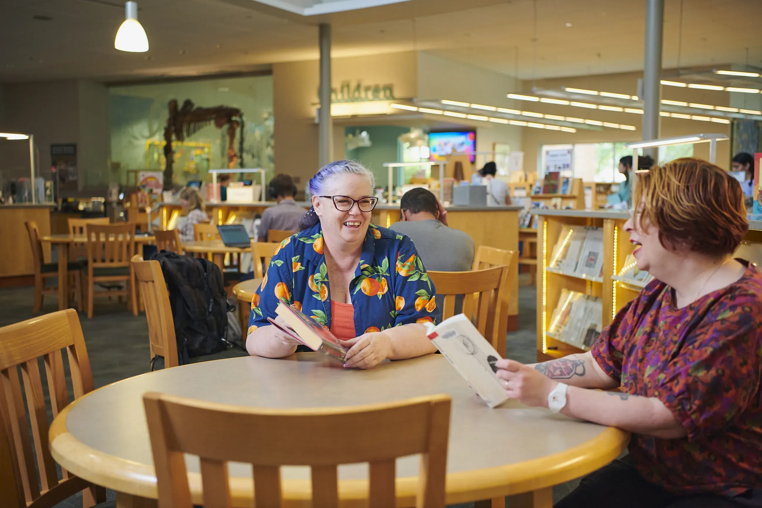 Two women holding books, seated at a table in a library.
