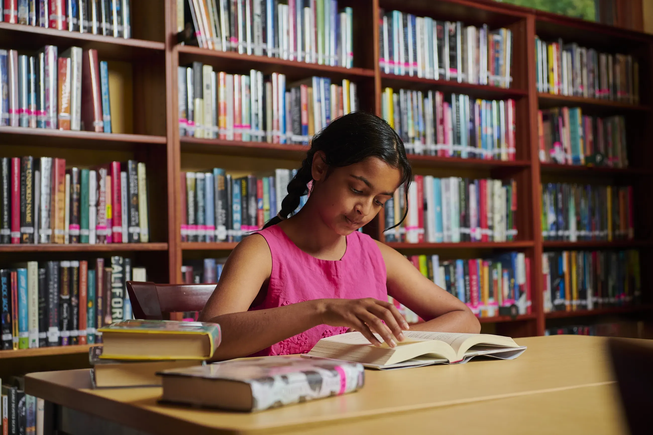 Young girl wearing pink tank top sitting and reading a book in a library.