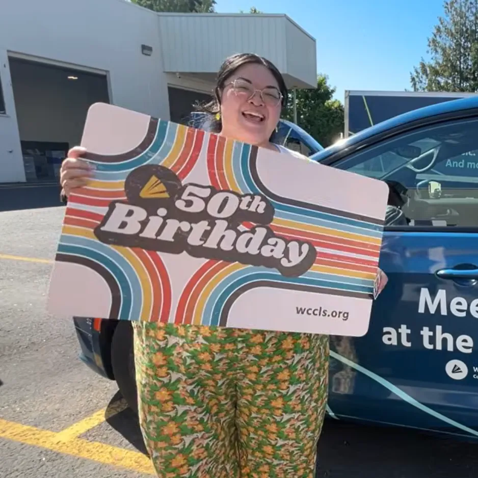 Woman with dark brown hair wearing glasses and colorful overalls. She is holding a giant WCCLS special edition library card. The card has rainbow stripes and the words 50th Birthday wccls.org on it.