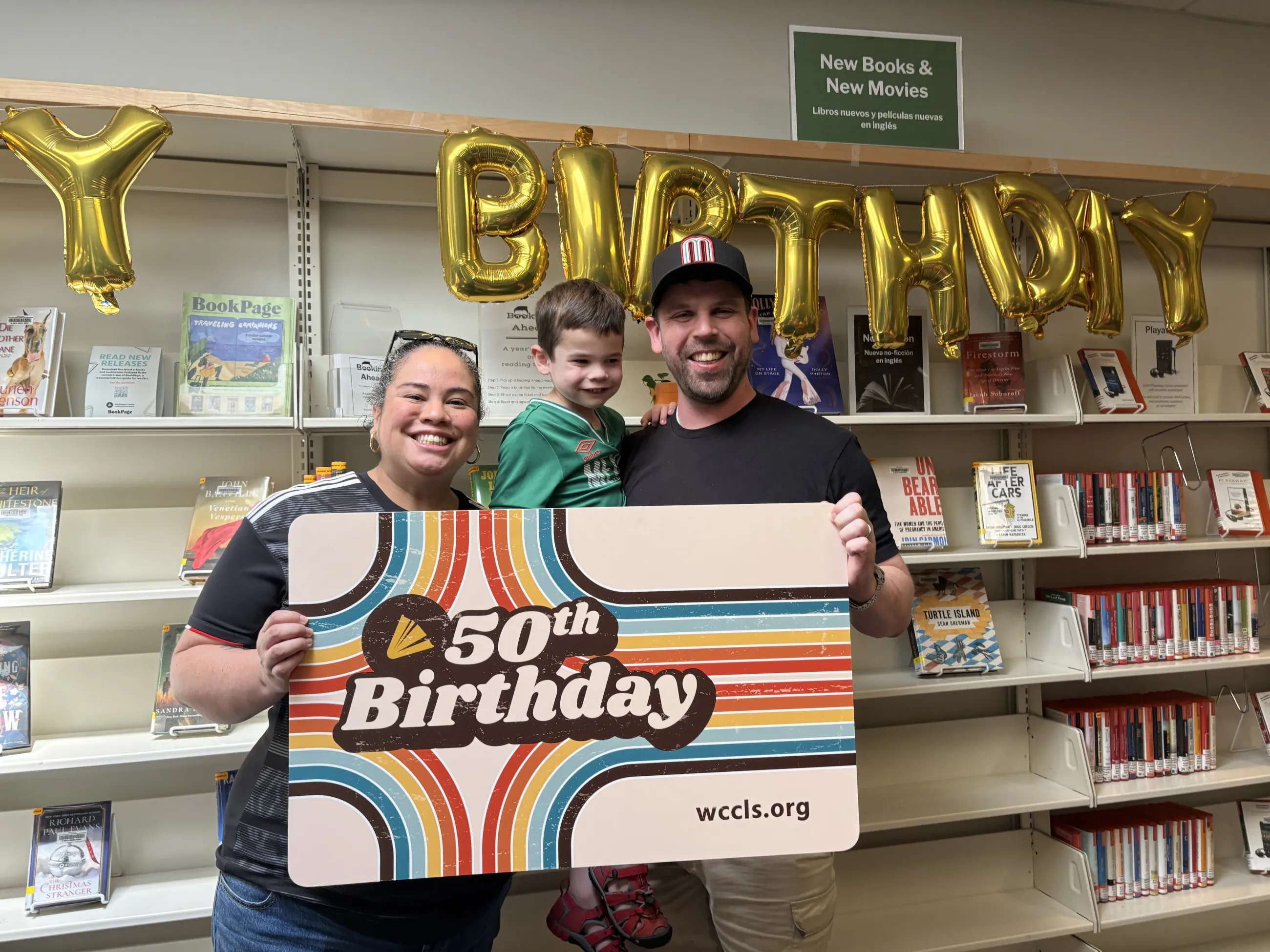 Mom, dad, and son posing in front of a gold birthday banner and holding the WCCLS 50th Birthday giant library card.