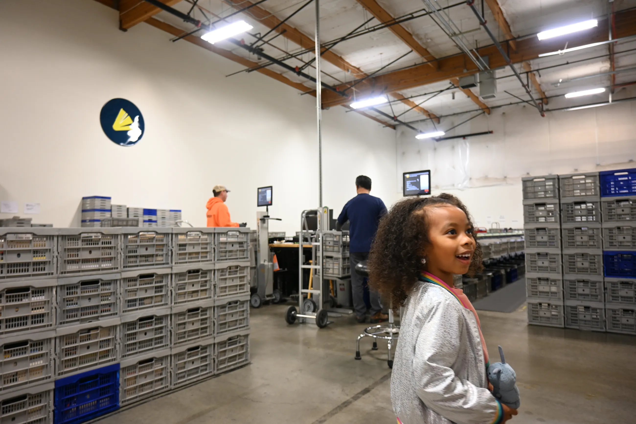 Young girl with brown curly hair and wearing a gray jacket smiles while touring the WCCLS warehouse.