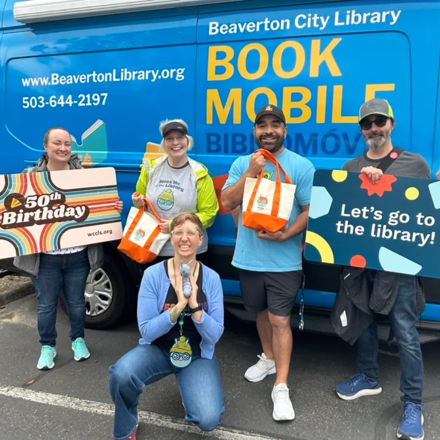 5 people pose in front of the Beaverton bookmobile holding giant library cards, mini totes, and Wallace the Book Bug.