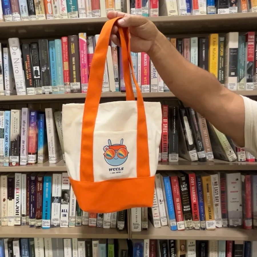 Hand holding up an orange and tan mini tote with a retro Wallace the Book Bug on the front pocket.