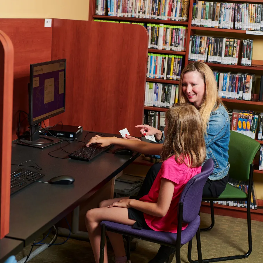 Woman and young girl sitting in front of desktop computer in library.