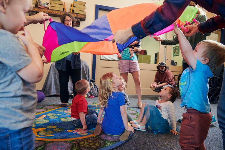 A group of young children, their parents, and library staff members, standing in a circle together and playing with a colorful parachute.