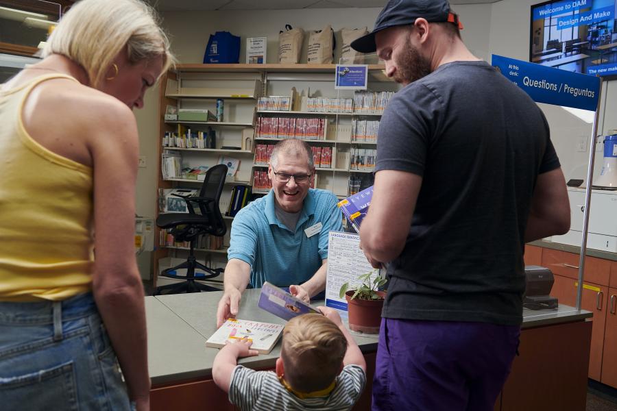 A mom, dad, and young son standing in front of the front desk and passing picture books to a library staff person who is smiling.