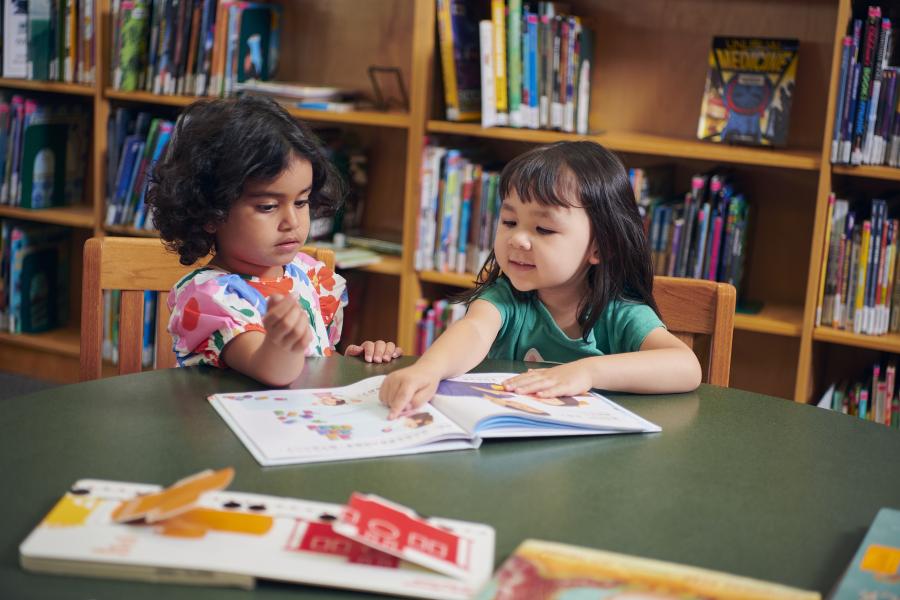 Two young girls sitting at a table and reading a picture book together in the library.