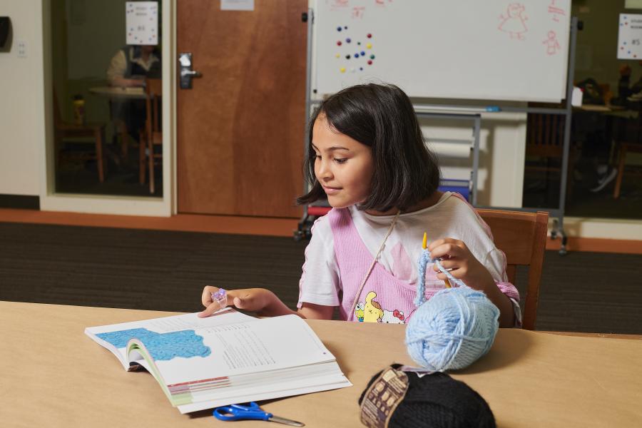 Young girl with short hair looking at crochet instruction book while holding a crochet needle and yarn.