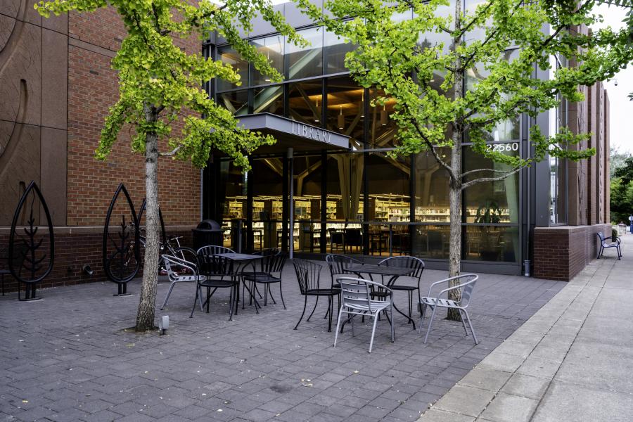 Exterior courtyard of the Sherwood Public Library that has two tables, an assortment of chairs, and two trees.